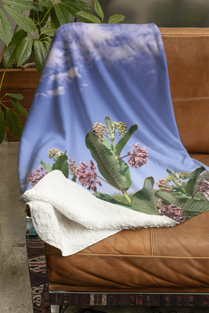 Couverture Lasclay imprimée de fleurs d’asclépiade commune sur fond de ciel bleu – posée sur un fauteuil.