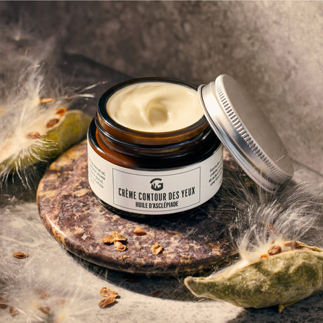 Pot ouvert de crème contour des yeux à l'huile d'asclépiade, posé sur une pierre avec des gousses et de la soie d'asclépiade, collaboration Gourmet Sauvage.