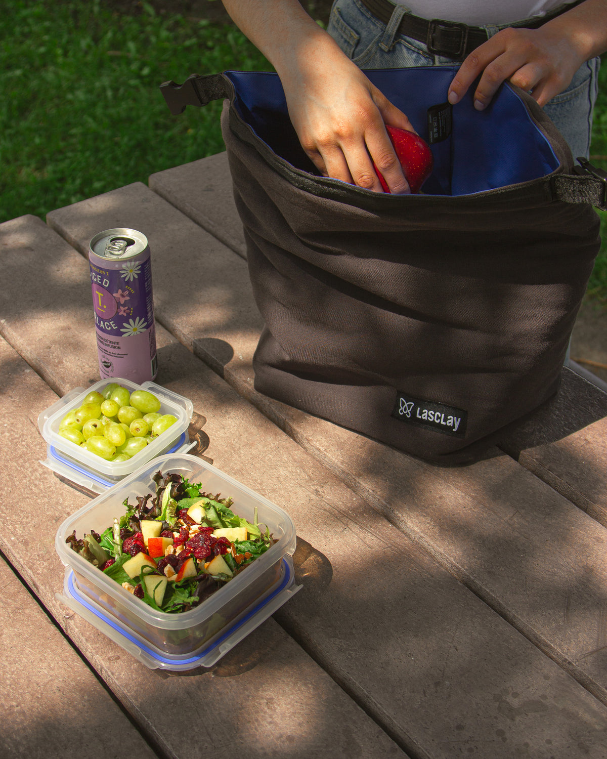 Sac à lunch isotherme Lasclay gris à doublure bleue posé sur une table de pique-nique, avec salade, raisins et une canette à côté.