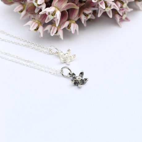 Deux colliers à pendentif fleur d'asclépiade en argent, l'un poli l'autre patiné, présentés devant des fleurs roses d'asclépiade.