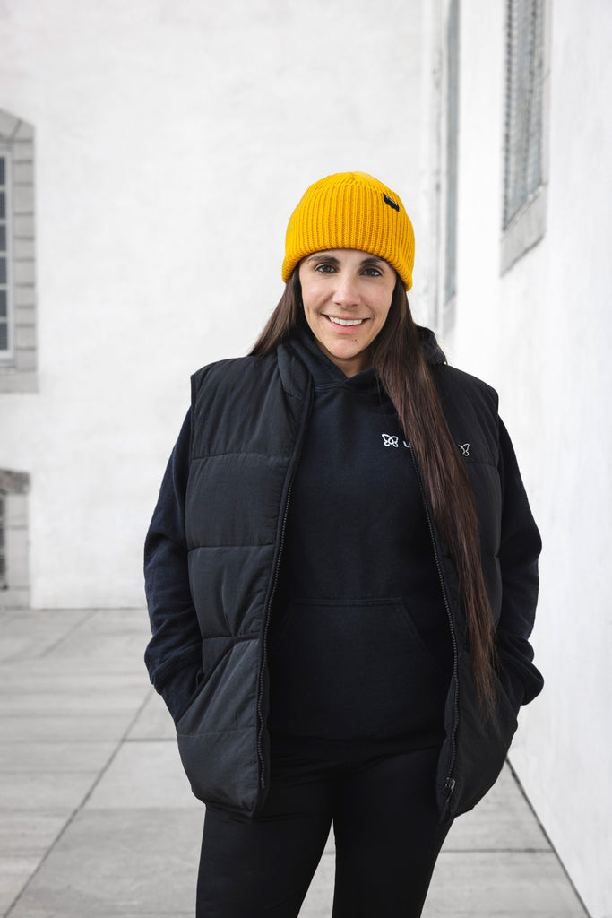 Femme souriante avec tuque jaune tricotée isolée d'asclépiade et veste sans manche noire Lasclay dans environnement urbain